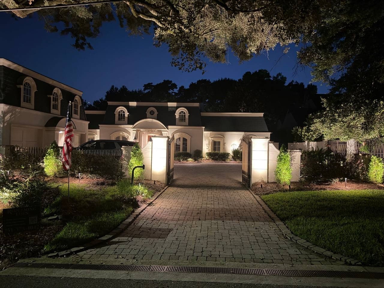 Gated stucco estate with a mansard roof, seen across a paver driveway lit between two illuminated gate piers, oak canopy overhead