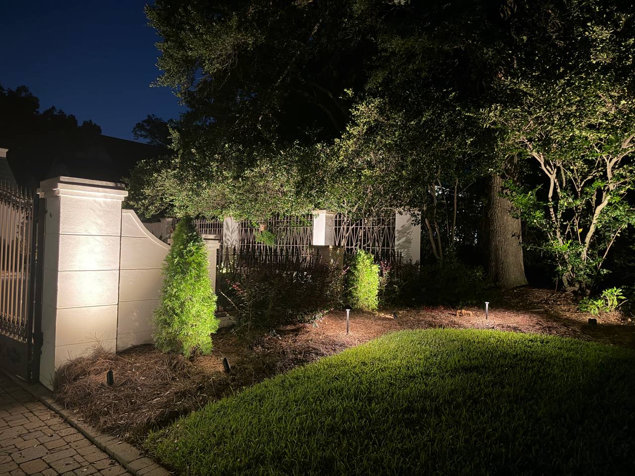 White stucco gate pier and iron fence lit by a wall-mounted floodlight beneath a large live oak canopy