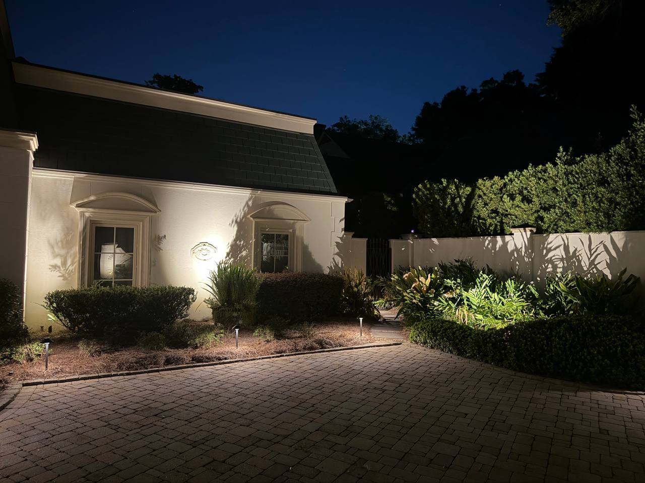 Side of the home with arched windows above a wide paver motor court and a lit foundation planting bed