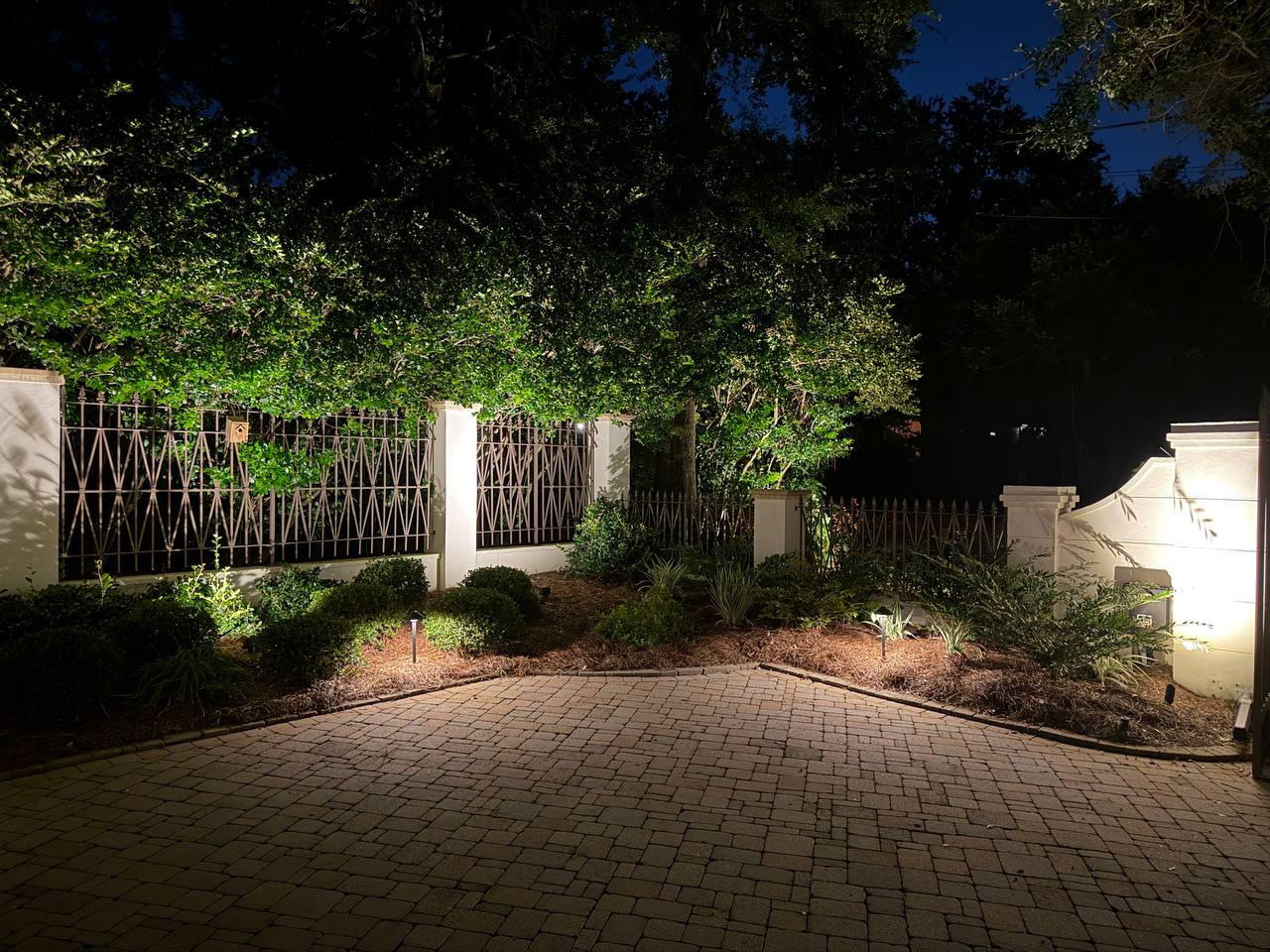 Decorative iron gate and stucco piers lit beneath a wide live oak, with foundation bed lighting along the drive