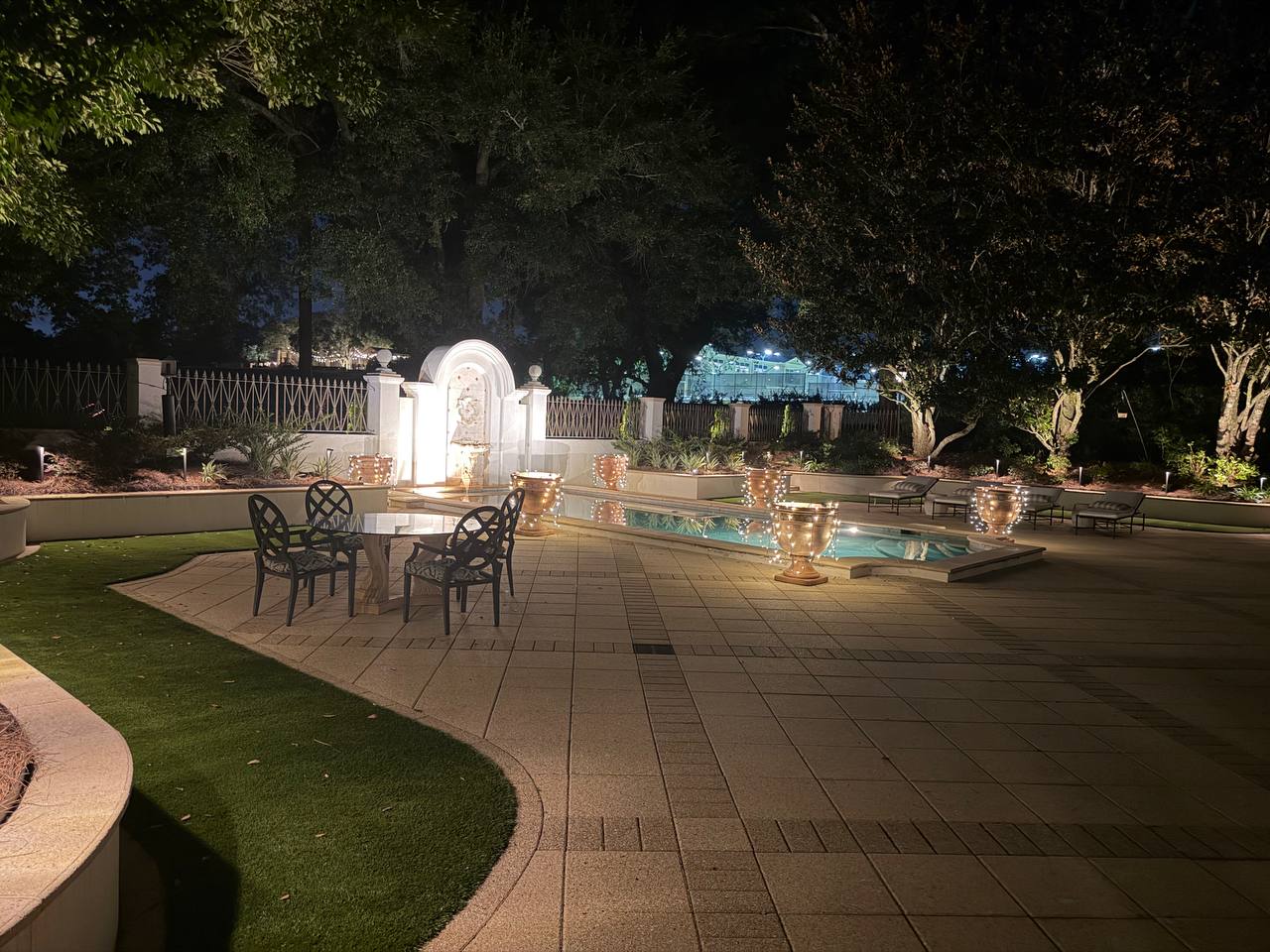 Wide pool terrace with a lit reflecting pool lined by string-light-wrapped urns, an iron dining table, an arched wall fountain, and lounge chairs under an oak canopy