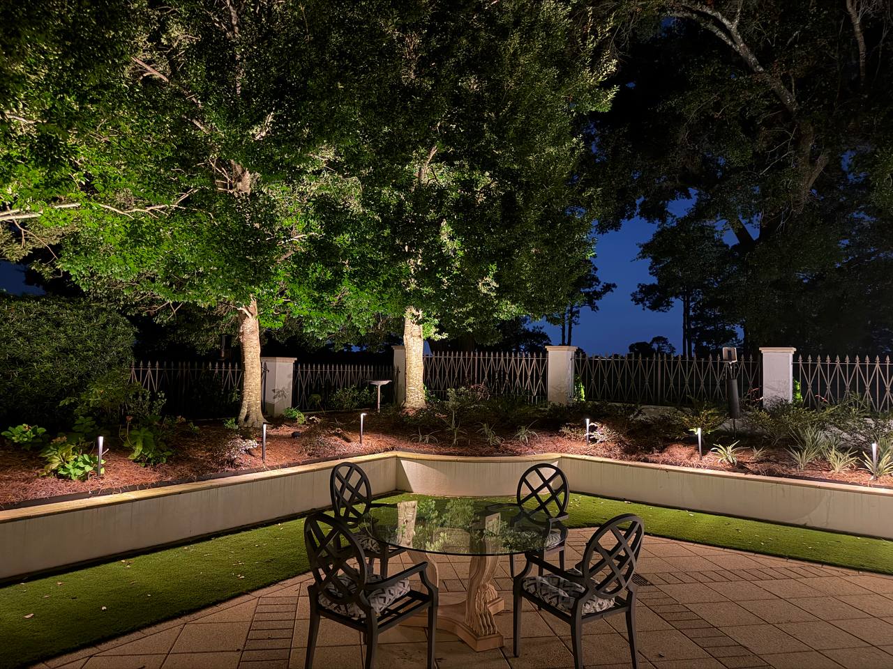 Glass-top iron dining table and chairs on a paver terrace, with path lights along a raised bed and two uplit tree trunks behind a picket fence